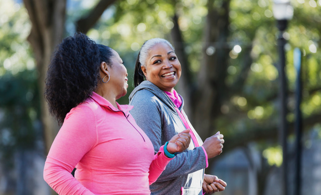 Two African-American women exercising together in the city, jogging or power walking, laughing and conversing. Buildings and trees are out of focus in the background. The one in pink is in her 60s and her friend is in her 50s.